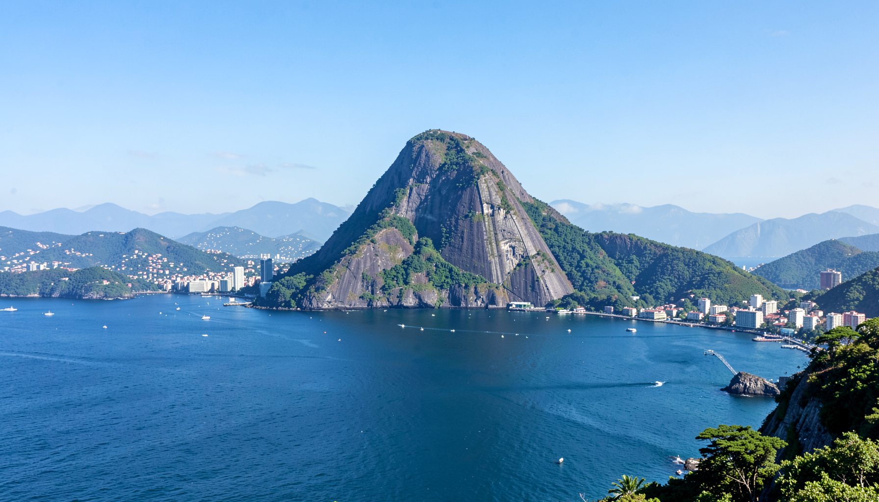 Iconic Sugarloaf Mountain rising above Guanabara Bay with Rio de Janeiro skyline and green hills in the background under a bright sky